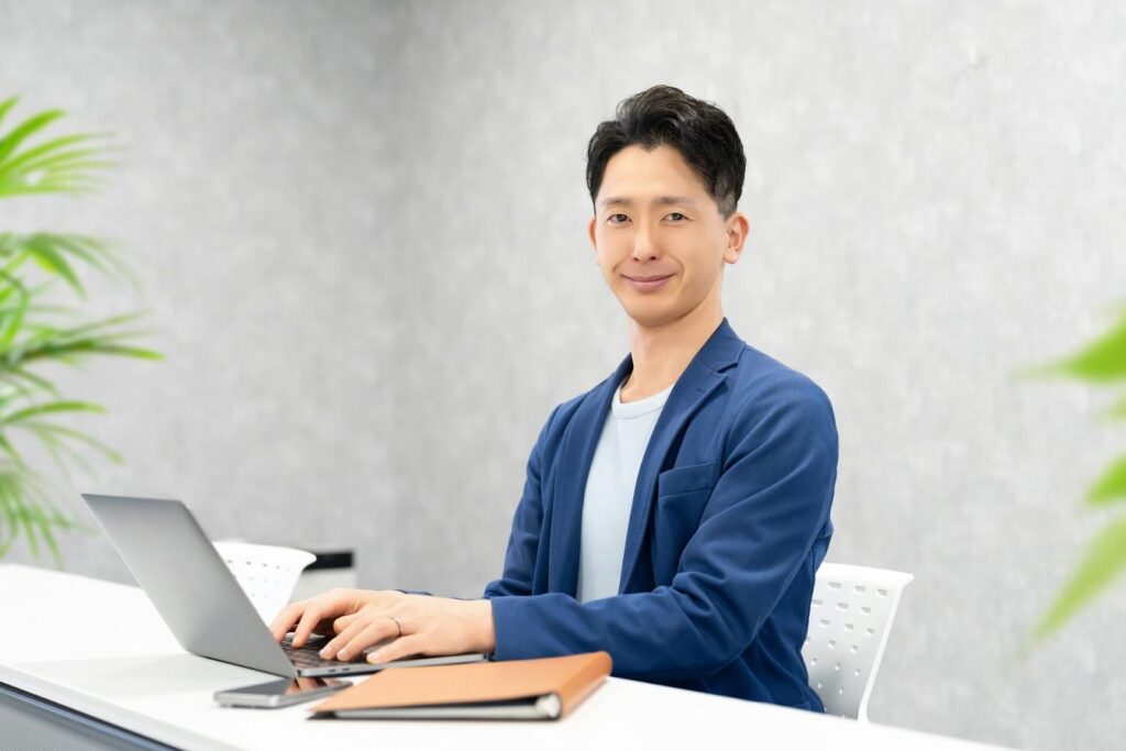 パソコンを打ちながらこちらを見る青いオフィスカジュアル服の男性