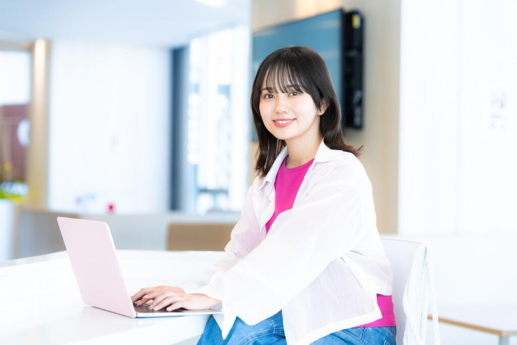 パソコンを作業しながらこちらを向いて微笑む白い私服の女性