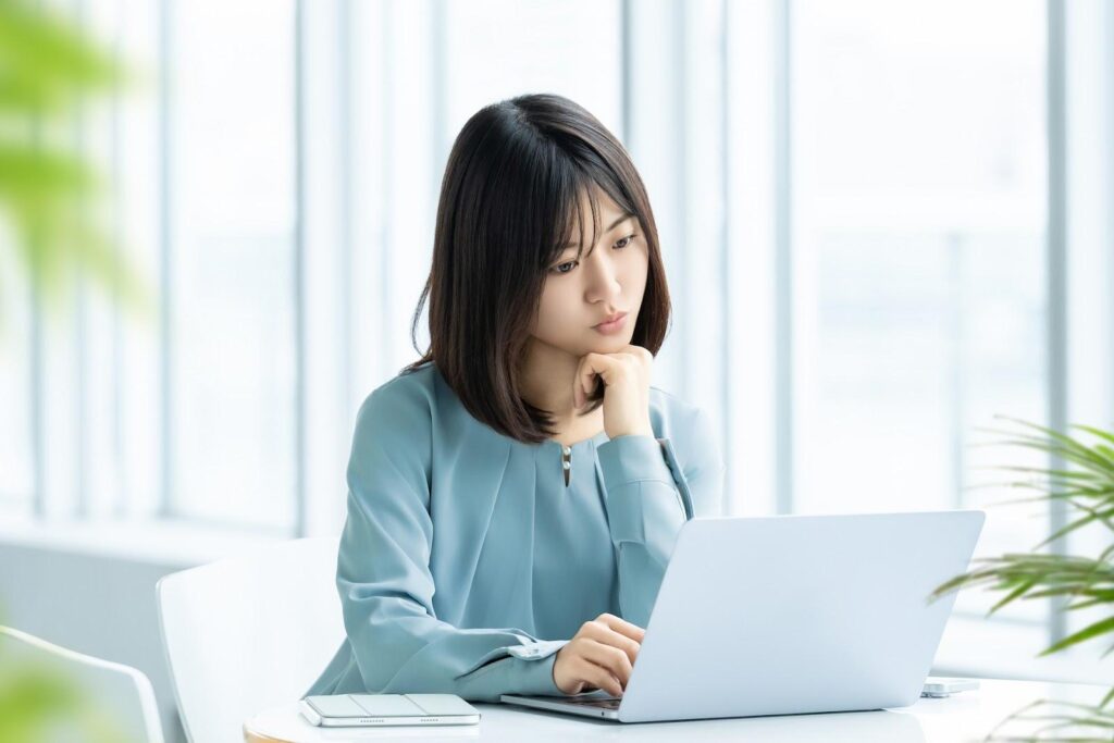 オフィスでパソコンを開きながら顎に手を当てて不思議そうな表情を浮かべる女性