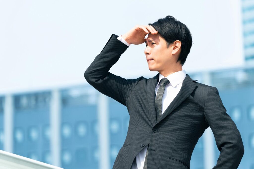 片手で日差しを遮りながら眩しそうに遠くを見つめるスーツ男性