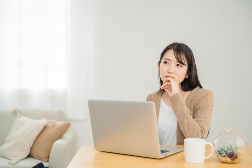 自宅でパソコンを開きながら顎に手を当てて悩む私服女性