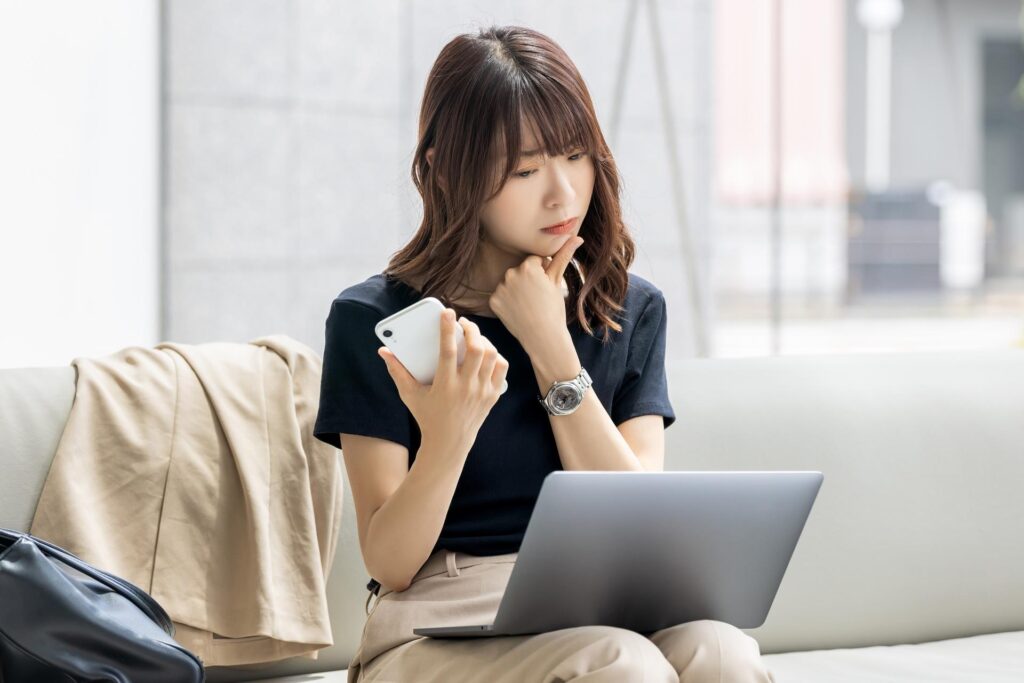 パソコンと携帯を眺めながら顎に指を当てて悩んでいる女性