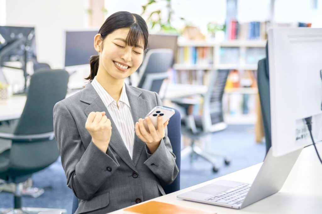 スマホとパソコンを見ながらガッツポーズするスーツの女性