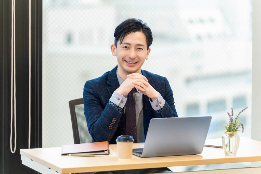 オフィスのデスクの前で上でを組んでこちらに向かって微笑む中年ビジネスマン