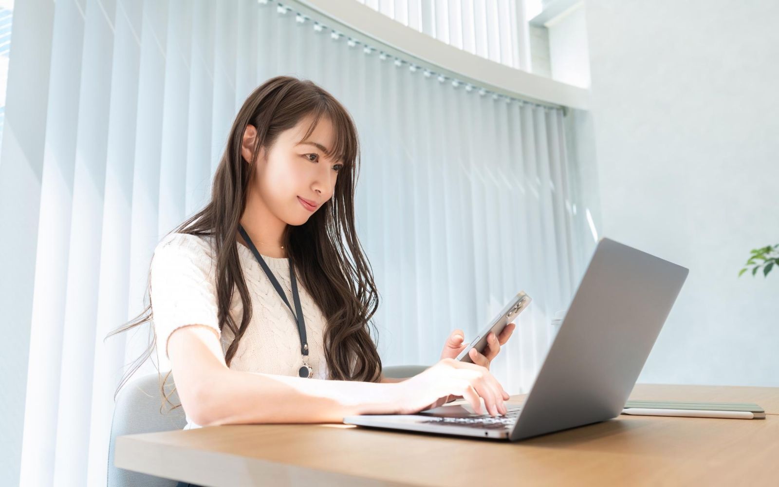 資料とスマートフォンを見ながらノートパソコンを使う女性
