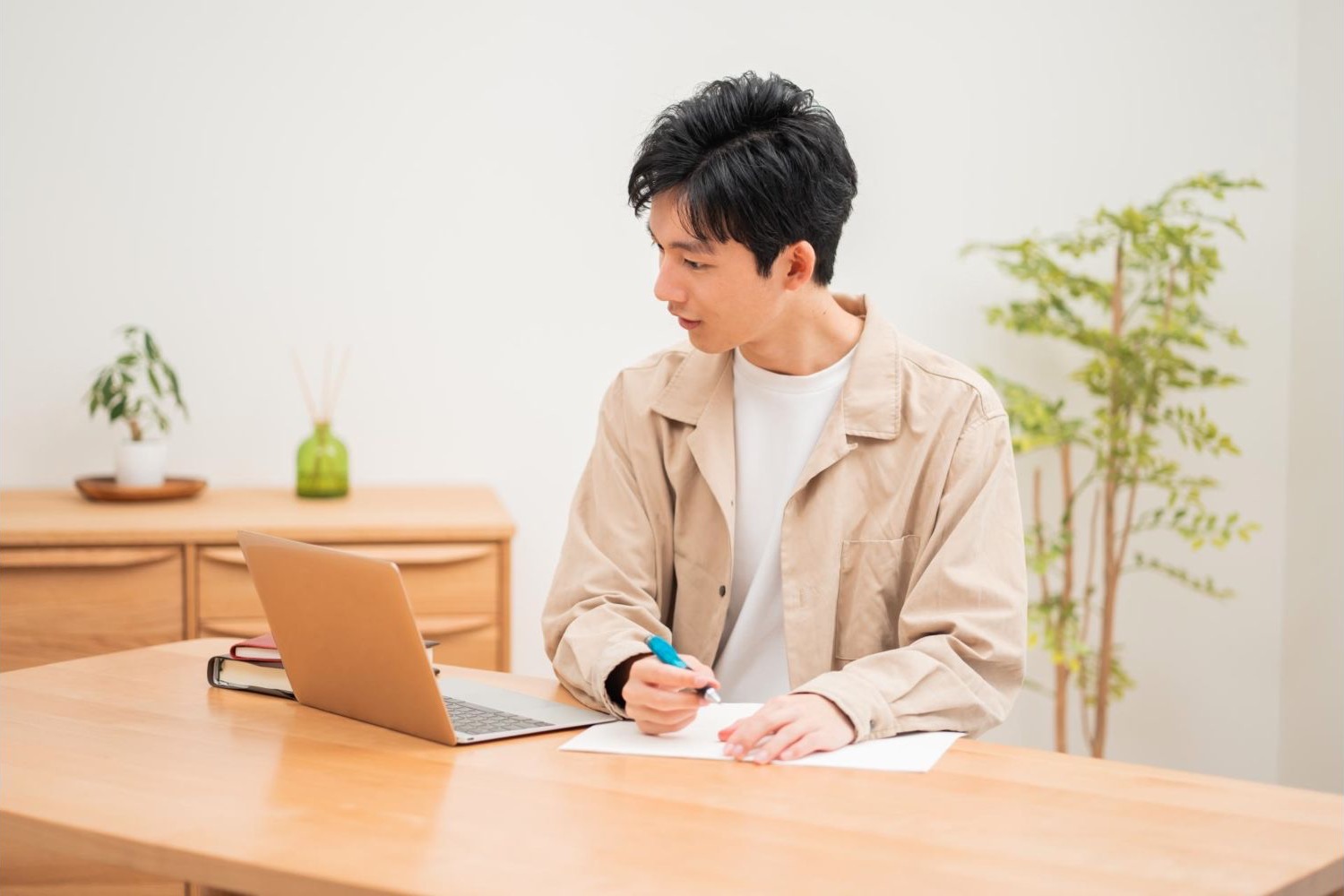 自宅の机でノートに書きながらノートパソコンを見る男性