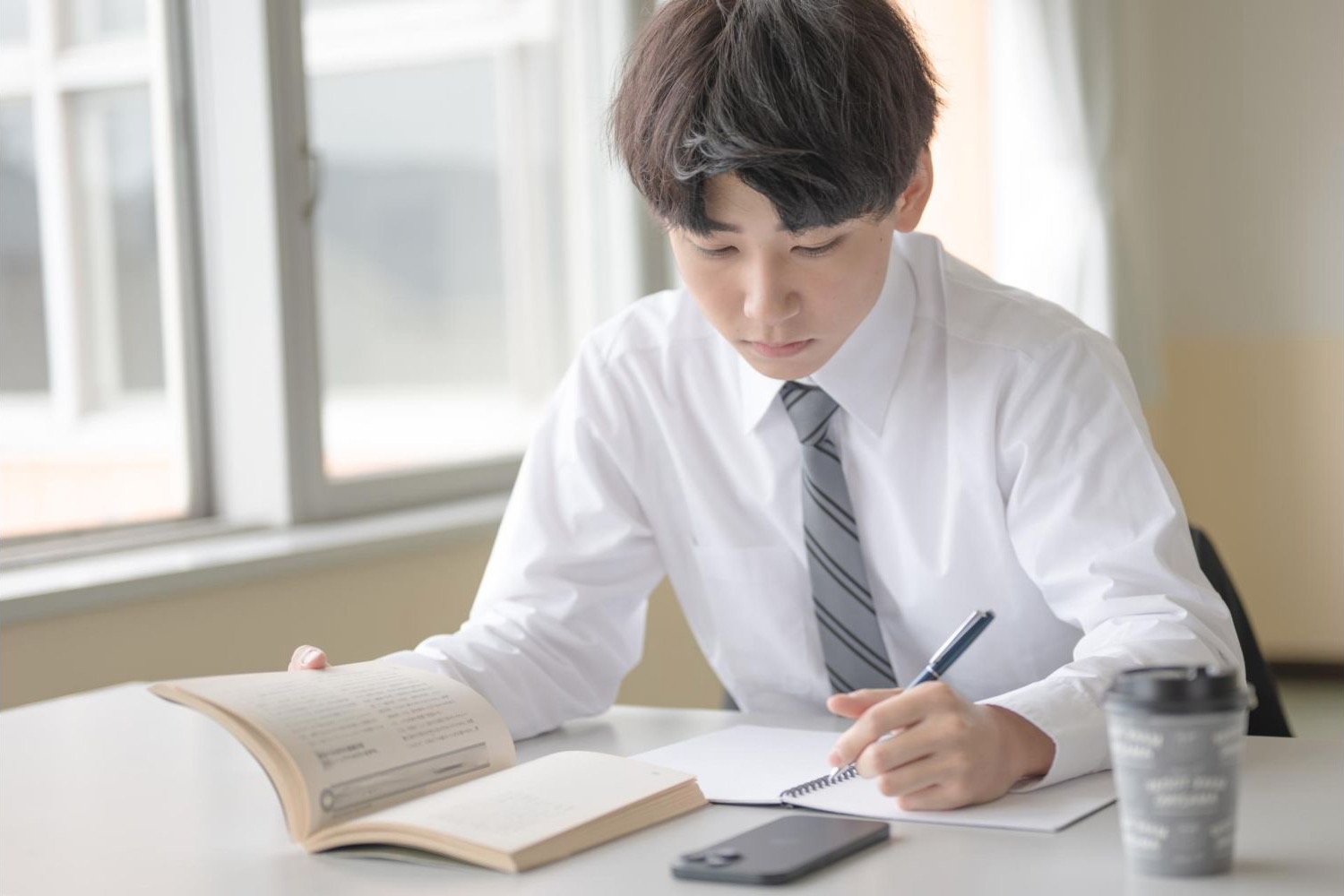 本とスマートフォンを置いた机でノートに書く男性