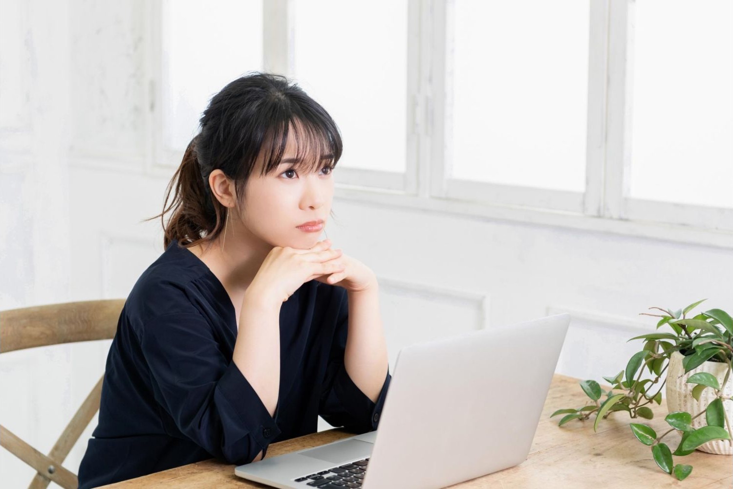 ノートパソコンの横で窓の外を見ながら考える女性