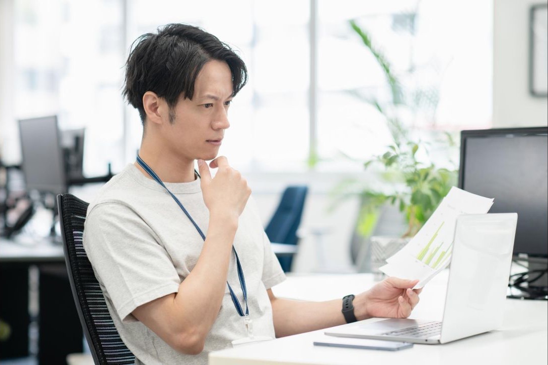 資料を手にノートパソコンを見ながら考える男性会社員